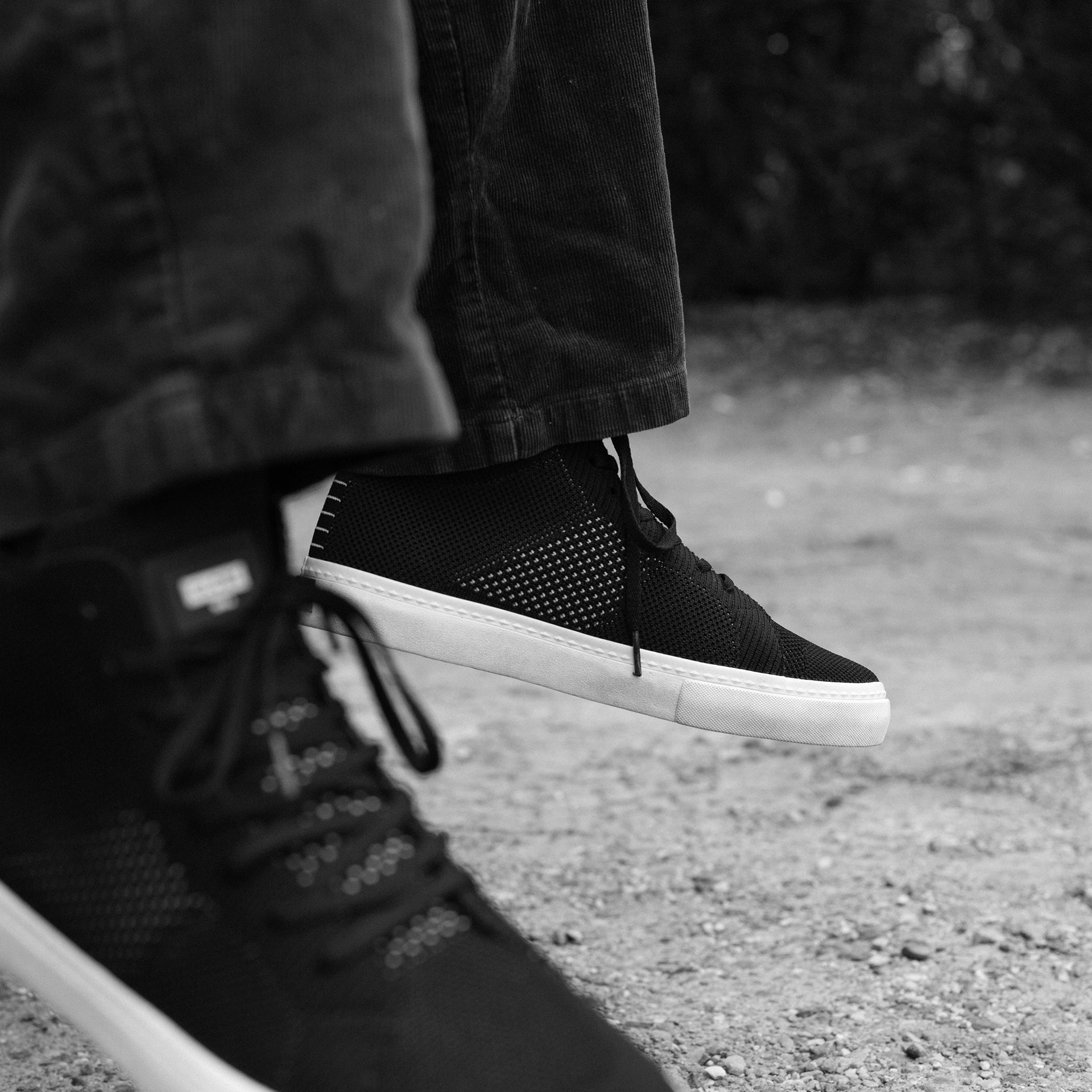 Black sneakers with white soles worn by a person on a gravel surface - Velocity 21